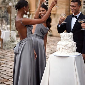 K&ouml;nnte beinhalten: Eine Feierszene mit Menschen, die Champagnergl&auml;ser erheben. Zwei Frauen tragen graue Kleider, eine mit Schleife. Ein Mann im Smoking ist dabei. Ein wei&szlig;er mehrst&ouml;ckiger Kuchen steht auf einem Tisch mit wei&szlig;em Tuch. Die Szene spielt sich im Freien ab.
