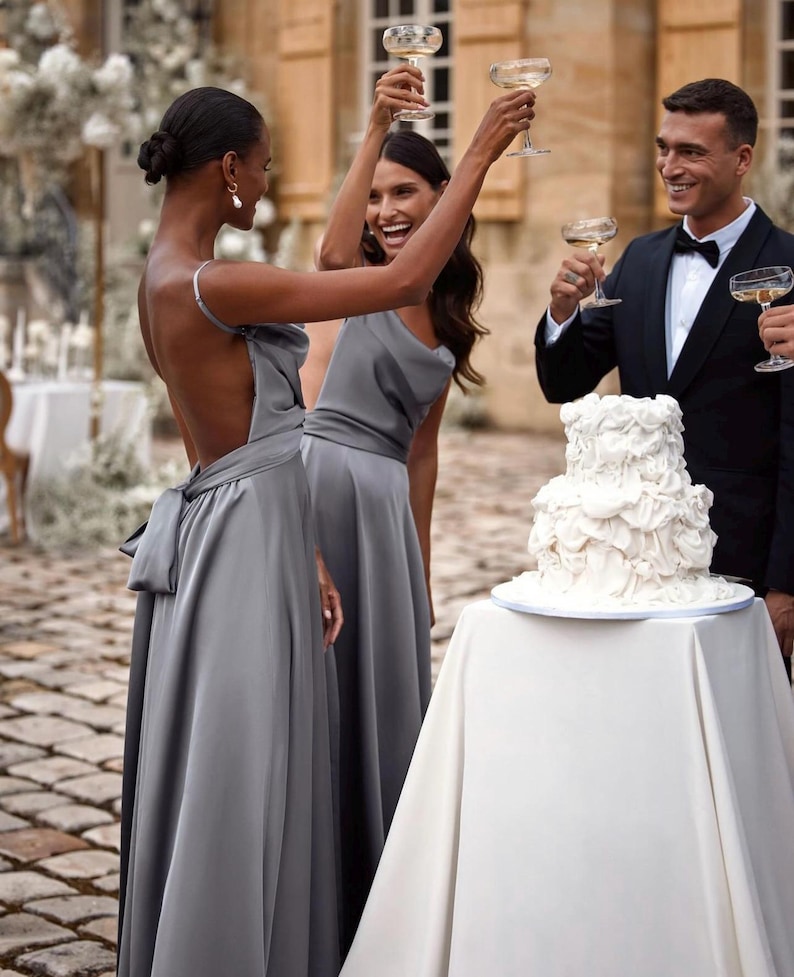 K&ouml;nnte beinhalten: Eine Feierszene mit Menschen, die Champagnergl&auml;ser erheben. Zwei Frauen tragen graue Kleider, eine mit Schleife. Ein Mann im Smoking ist dabei. Ein wei&szlig;er mehrst&ouml;ckiger Kuchen steht auf einem Tisch mit wei&szlig;em Tuch. Die Szene spielt sich im Freien ab.