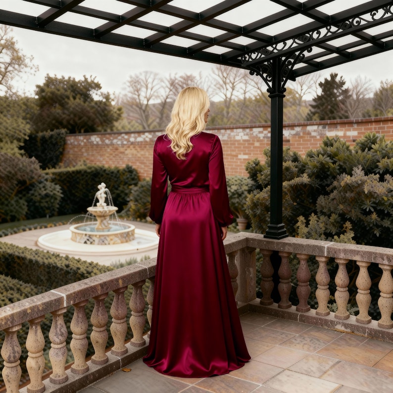 May include: A long, flowing burgundy satin robe with long sleeves and a belted waist. The robe is worn outdoors on a stone patio with a decorative pergola overhead. A fountain and manicured garden are in the background.