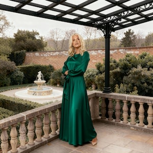 May include: A long, emerald green satin dress with long sleeves and a matching waist tie. The dress is worn with nude heels. The background features a garden with a fountain and a stone balustrade.