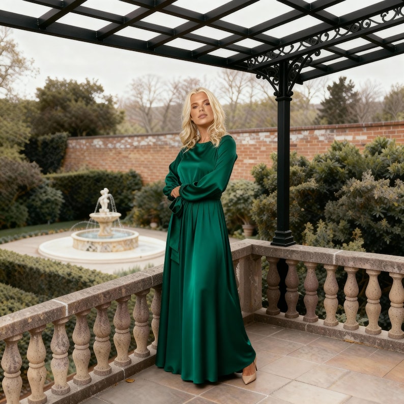 May include: A long, emerald green satin dress with long sleeves and a matching waist tie. The dress is worn with nude heels. The background features a garden with a fountain and a stone balustrade.