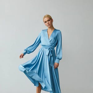 Puede incluir: Vestido cruzado de sat&eacute;n azul claro con mangas largas y cintur&oacute;n anudado. El vestido tiene un escote en V y una falda fluida. La modelo lleva tacones blancos. El vestido es de largo midi.