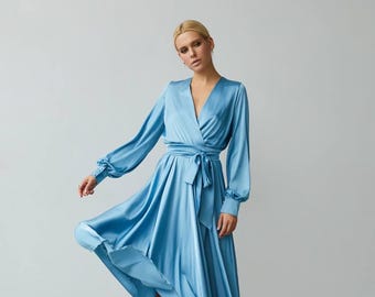 May include: A light blue satin wrap dress with long sleeves and a V-neck. The dress features a waist tie and a flowing skirt. The model is wearing white heels. The dress is knee-length.