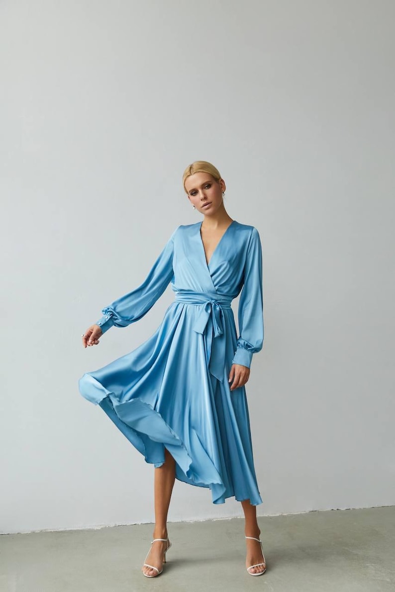 Puede incluir: Vestido cruzado de sat&eacute;n azul claro con mangas largas y cintur&oacute;n anudado. El vestido tiene un escote en V y una falda fluida. La modelo lleva tacones blancos. El vestido es de largo midi.