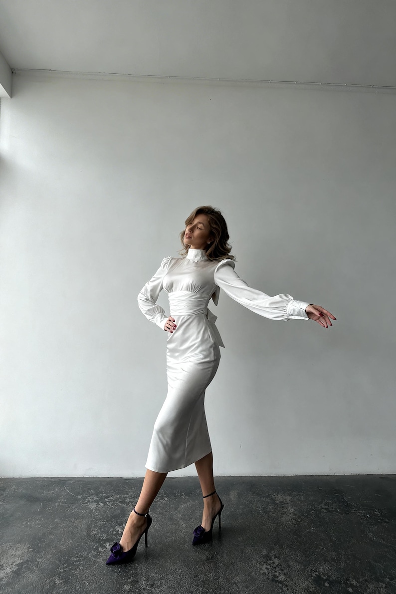 May include: A white satin midi dress featuring a high neckline, long sleeves with buttoned cuffs, and a fitted bodice with a waist tie. The dress has a smooth, lustrous finish and a straight skirt that falls below the knees, paired with dark heels.