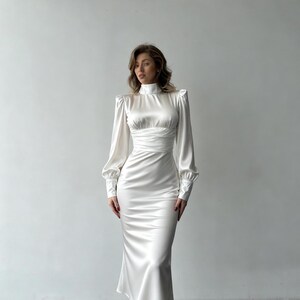 May include: A floor-length white satin dress featuring a high neckline, long sleeves with buttoned cuffs, and a gathered, ruched waistline. The garment has a fitted bodice and a flowing skirt that extends to the lower calf, crafted from a smooth, reflective fabric.
