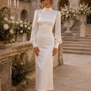 May include: A floor-length ivory satin dress featuring a high mock neckline, long sleeves with gathered shoulders and buttoned cuffs, and a fitted waistline. The garment has a smooth, lustrous finish and a straight skirt silhouette.