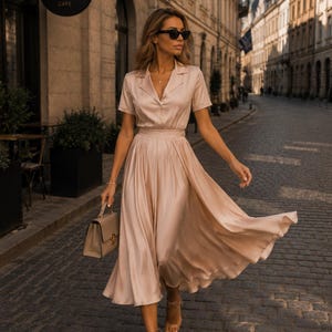 May include: A champagne-colored midi dress with a collared neckline, short sleeves, and a button-down bodice. The skirt is pleated and flows outward. A matching structured handbag with a top handle is held in one hand. The outfit is paired with clear strappy sandals and dark-framed sunglasses.