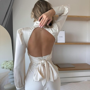May include: A long-sleeved cream-colored satin dress featuring a high neckline, a large open back design, and a prominent fabric tie closure at the waist. The garment has structured shoulders and fitted cuffs, showcasing a smooth, lustrous finish.