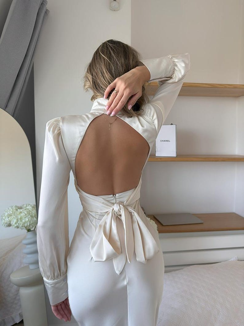 May include: A long-sleeved cream-colored satin dress featuring a high neckline, a large open back design, and a prominent fabric tie closure at the waist. The garment has structured shoulders and fitted cuffs, showcasing a smooth, lustrous finish.