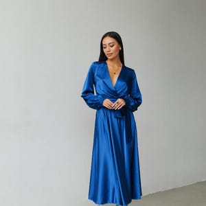 Puede incluir: Un vestido largo y fluido de sat&eacute;n azul real con mangas largas y cintur&oacute;n a juego. El vestido tiene un escote en V y llega hasta la pantorrilla. La modelo lleva sandalias blancas con tiras. Es una prenda de vestir formal.