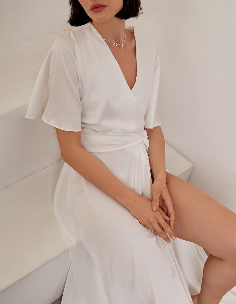 May include: A white wrap dress with short, flared sleeves and a V-neck. The dress is tied at the waist and has a flowing skirt. A delicate silver necklace with small pendants is worn with the dress.