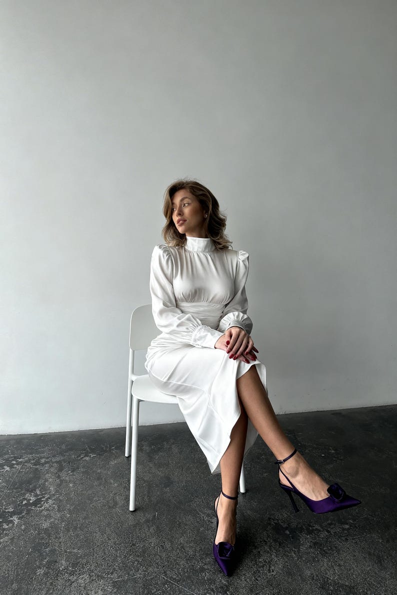 May include: A white satin midi dress featuring a high mock neckline, long sleeves with gathered cuffs, and a fitted waistline. The dress has a smooth, lustrous finish and a straight skirt that falls below the knees, paired with dark purple pointed heels.