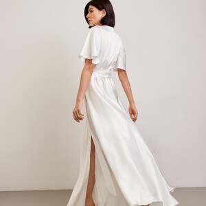 May include: A white satin maxi dress with a high slit and short, flutter sleeves. The dress is cinched at the waist with a matching belt. The model is wearing beige heels. The dress is flowing and elegant.