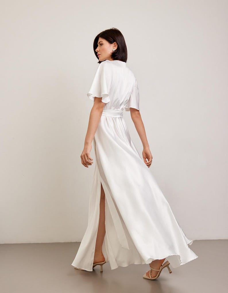 May include: A white satin maxi dress with a high slit and short, flutter sleeves. The dress is cinched at the waist with a matching belt. The model is wearing beige heels. The dress is flowing and elegant.