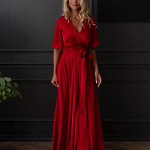 May include: A full-length, red wrap dress with short sleeves and a matching waist tie. The dress has a V-neckline and a flowing skirt. The woman in the image is wearing the dress and has blonde hair.