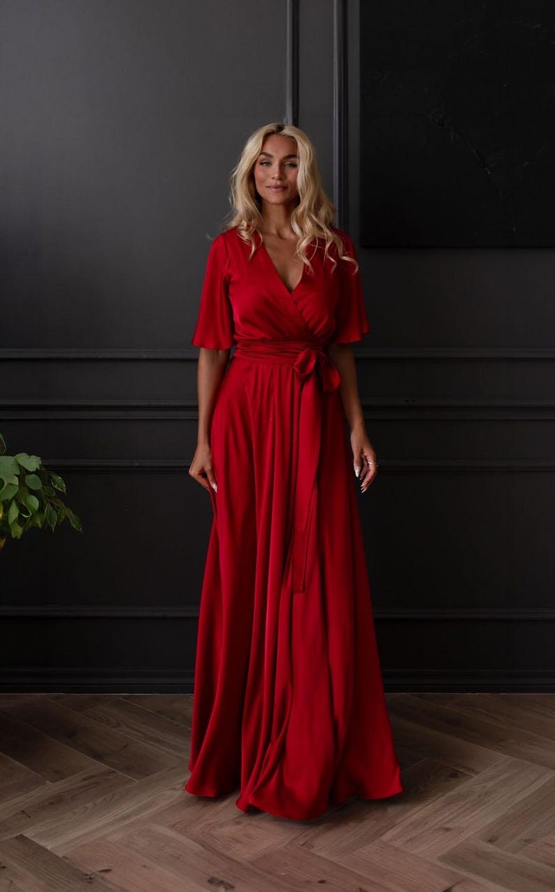 May include: A full-length, red wrap dress with short sleeves and a matching waist tie. The dress has a V-neckline and a flowing skirt. The woman in the image is wearing the dress and has blonde hair.