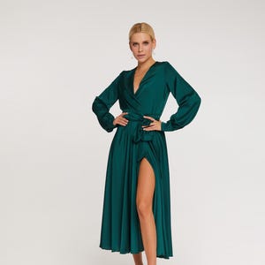 Puede incluir: Un vestido largo cruzado de color verde esmeralda con mangas largas y una abertura alta. El vestido est&aacute; ce&ntilde;ido a la cintura con un cintur&oacute;n de tela a juego. La modelo lleva tacones blancos con tiras. El vestido est&aacute; hecho de una tela brillante y fluida.