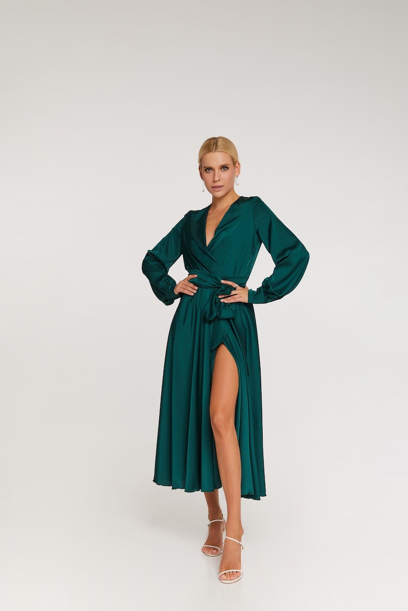 Puede incluir: Un vestido largo cruzado de color verde esmeralda con mangas largas y una abertura alta. El vestido est&aacute; ce&ntilde;ido a la cintura con un cintur&oacute;n de tela a juego. La modelo lleva tacones blancos con tiras. El vestido est&aacute; hecho de una tela brillante y fluida.