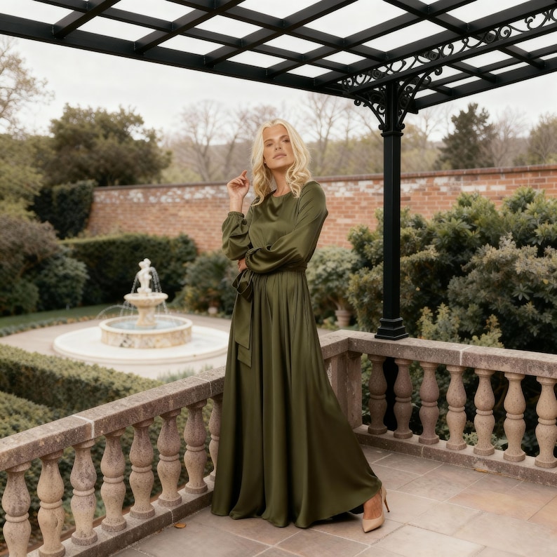 May include: An olive green maxi dress with long sleeves and a matching belt. The dress features a round neckline and is made of a silky fabric. The model is wearing nude heels. The background includes a stone balcony, a fountain, and green foliage.