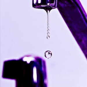 Puede incluir: Primer plano de un grifo plateado con una sola gota de agua colgando del caño. El grifo está sobre un fondo morado.
