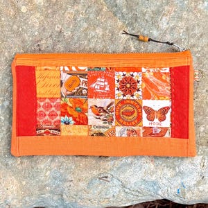 May include: An orange patchwork zippered pouch with a variety of vintage-style images. The pouch features a mix of orange, red, and cream-colored squares with images of flowers, birds, and text. The zipper pull has a wooden bead.
