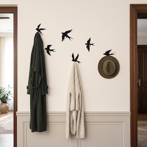 Op de afbeelding: Een wanddecoratie met zwarte zwaluw silhouetten in vlucht. Een donkergroene badjas en een crèmekleurige trui hangen aan de muur. Een groene hoed rust op een haak. De muur is lichtbeige geschilderd.