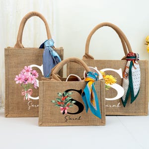 May include: Three jute tote bags with brown handles, each featuring a floral or botanical design and the letter "S". The bags have decorative scarves tied to the handles. The word "Sarah" is printed on two of the bags.