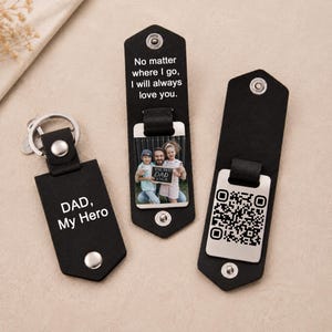 Peut inclure: Trois porte-clés en cuir noir avec des boutons-pression en métal argenté. L'un porte l'inscription 'DAD, My Hero'. Un autre affiche une photo d'un homme et deux enfants tenant une pancarte 'THE BEST DAD EVER' avec le texte 'No matter where I go, I will always love you.' Le troisième présente un code QR.