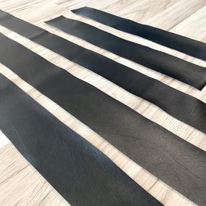 Peut inclure: Plusieurs longues bandes de cuir noir sont disposées en diagonale sur une surface en bois clair. Les bandes sont espacées de manière régulière, créant un motif de lignes noires et marron clair alternées. Le cuir est légèrement texturé.