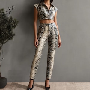 Puede incluir: Un conjunto de dos piezas con estampado de piel de serpiente. El conjunto incluye un top corto con cuello y mangas cortas y pantalones de cintura alta a juego. El atuendo es en tonos grises, beige y negro. La modelo lleva tacones negros.