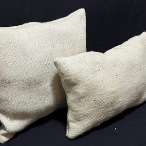 May include: Two off-white, textured throw pillows. The pillows are rectangular with a slightly irregular shape, suggesting a handmade quality. The fabric has a woven appearance, and the pillows are placed against a dark background.