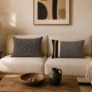Puede incluir: Dos cojines rectangulares colocados sobre un sofá beige. El cojín izquierdo presenta un patrón de puntos en blanco y negro. El cojín derecho muestra un diseño de cuadrícula geométrica en blanco y negro con una franja vertical en zigzag.