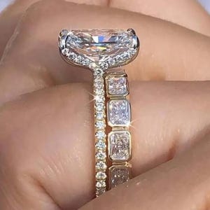 Puede incluir: Un juego de anillos de diamantes de dos piezas. El anillo de compromiso presenta un diamante grande de corte rectangular con un engaste de halo. La banda está adornada con diamantes más pequeños. La banda a juego tiene diamantes rectangulares y una banda dorada.