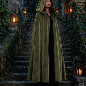 May include: A floor-length olive green velvet cloak featuring a deep hood and a metallic clasp closure at the neck. The garment is worn over a simple black dress, with the fabric draping in soft, vertical folds down to the hemline.