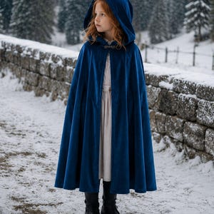 May include: A deep blue velvet hooded cloak featuring a metallic clasp at the neckline, worn over a light-colored dress and black boots. The cloak has a full-length design with a structured hood and soft fabric texture that drapes down to the knees.