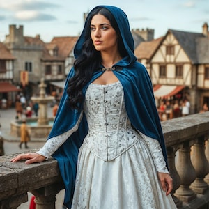 May include: A woman wearing a medieval-style gown with a white bodice and skirt, adorned with floral patterns. A hooded, dark blue velvet cape is draped over the shoulders. The background features a European town with stone buildings and a fountain.