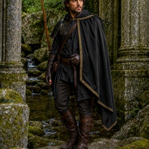 May include: A black hooded cloak with gold trim worn over a dark tunic and trousers. The outfit includes brown leather gloves, a brown leather belt with a pouch, and tall brown leather boots. A wooden bow is carried over the shoulder.