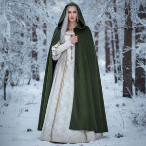 May include: A woman wearing a long, white dress with gold trim and a dark green hooded cloak. The dress has long sleeves and a high neckline. The cloak is buttoned at the neck. The background is a snowy forest.