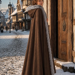 May include: A floor-length brown cloak featuring a hood trimmed with grey faux fur. The garment has a long, flowing silhouette that extends to the ground, with a continuous grey faux fur border running along the front opening and around the hood.