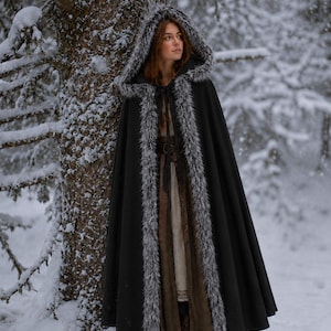 May include: A floor-length black cloak featuring a hood and front opening trimmed with grey and white faux fur. The garment is secured at the neck with thin black ties and is worn over a long, neutral-toned dress with a brown belt and dark boots.