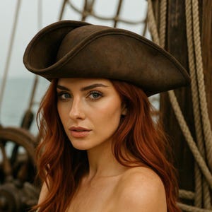 May include: A brown pirate hat with a wide brim. The hat is made of a soft material and has a slightly distressed look. The hat is worn by a person with long red hair. The background includes a ship's wheel and ropes, suggesting a nautical theme.