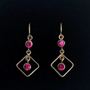 May include: A pair of gold-coloured drop earrings. Each earring has three pink gemstones and a diamond-shaped frame. The gemstones are round and set in gold-coloured metal. The earrings hang from a hook.