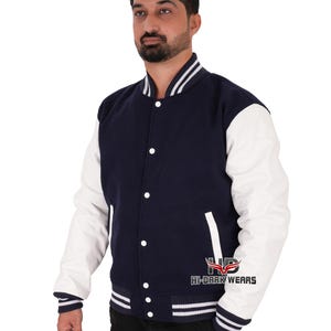 May include: A navy blue and white varsity jacket with a white leather sleeve. The jacket has a button closure and a ribbed collar, cuffs, and hem. The jacket has a white stripe on the collar, cuffs, and hem. The jacket has a pocket on the left side. The jacket has the text "HI-DARK WEARS" on the left side.