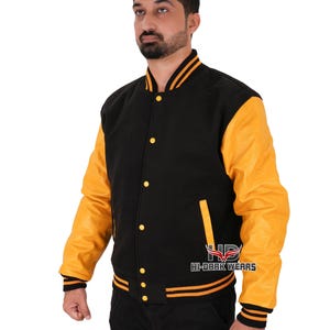 Puede incluir: Una chaqueta de béisbol negra y amarilla con cierre de presión y ribetes a rayas. La chaqueta tiene una manga de cuero amarilla y un cuerpo de lana negra.