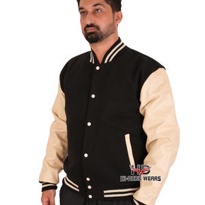 Pode incluir: Uma jaqueta de beisebol preta com detalhes em branco e preto listrados ao redor do colarinho, punhos e bainha. As mangas são de couro creme. A jaqueta tem fecho de botões e um bolso no lado esquerdo. O texto "HI-DARK WEARS" está impresso na parte inferior direita da jaqueta.