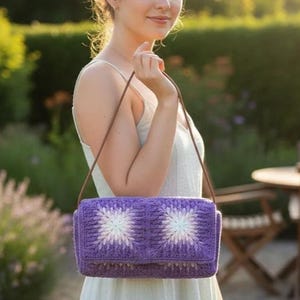 May include: A purple crochet shoulder bag with a floral design. The bag features a rectangular shape with a flap closure and a brown strap. The floral pattern is white and light purple, and the bag is held by a person.