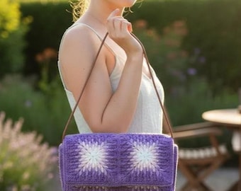 May include: A purple crochet shoulder bag with a floral design. The bag features a rectangular shape with a flap closure and a brown strap. The floral pattern is white and light purple, and the bag is held by a person.