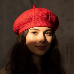 以下が含まれることがあります： 鮮やかな赤色の、かぎ針編みのベレー帽。ベレー帽は、丸いクラウンと上部の小さな茎を備えたクラシックなスタイルです。帽子は質感のある糸でできており、色は鮮やかで無地の赤です。ベレー帽は、黒髪の人が着用しています。