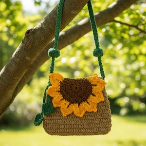 Op de afbeelding: Gehaakte schoudertas met een zonnebloemontwerp. De tas heeft een bruine body, een geel en bruin zonnebloem applicatie en een groene band. De tas hangt aan een tak, met een vage groene achtergrond.